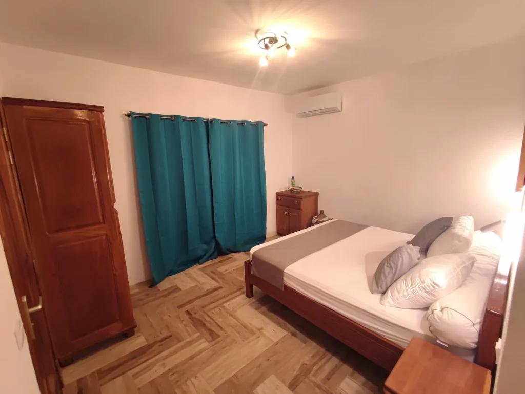 Vue d'ensemble de la chambre numéro 2, située au rez-de-chaussée de la première villa, dotée d'un lit King size, d'une literie confortable, d'une douche à l'italienne, d'un meuble salle de bain avec vasque et miroir, d'un cabinet de toilette séparé, d'une commode, d'une grande armoire, de luminaires et rideaux occultant bleu azur.