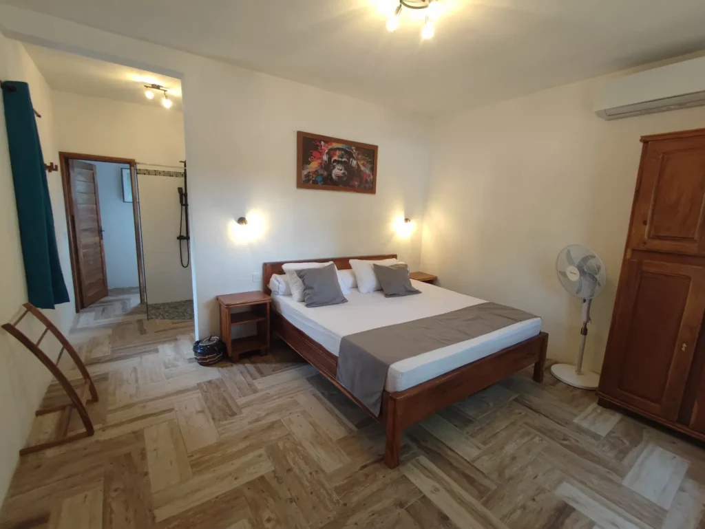 Le Campement Houéfa compte deux villas, avec chacune 3 chambres. Photo de la chambre numéro 4 avec son lit King size drapé de blanc, d'un chemin de lit et de coussins d'apparat de couleur gris clair, rappelant avec subtilité les tons de l'espace douche et son imposant tableau surplombant le lit.
