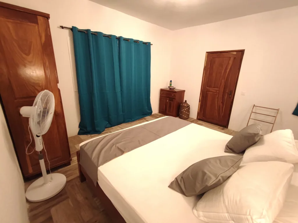 Vue d'ensemble de la chambre numéro 4, située au rez-de-chaussée de la seconde villa, dotée d'un lit King size, d'une literie confortable, d'une douche à l'italienne, d'un meuble salle de bain avec vasque et miroir, d'un cabinet de toilette séparé, d'une commode, d'une grande armoire, de luminaires et rideaux occultant bleu azur.