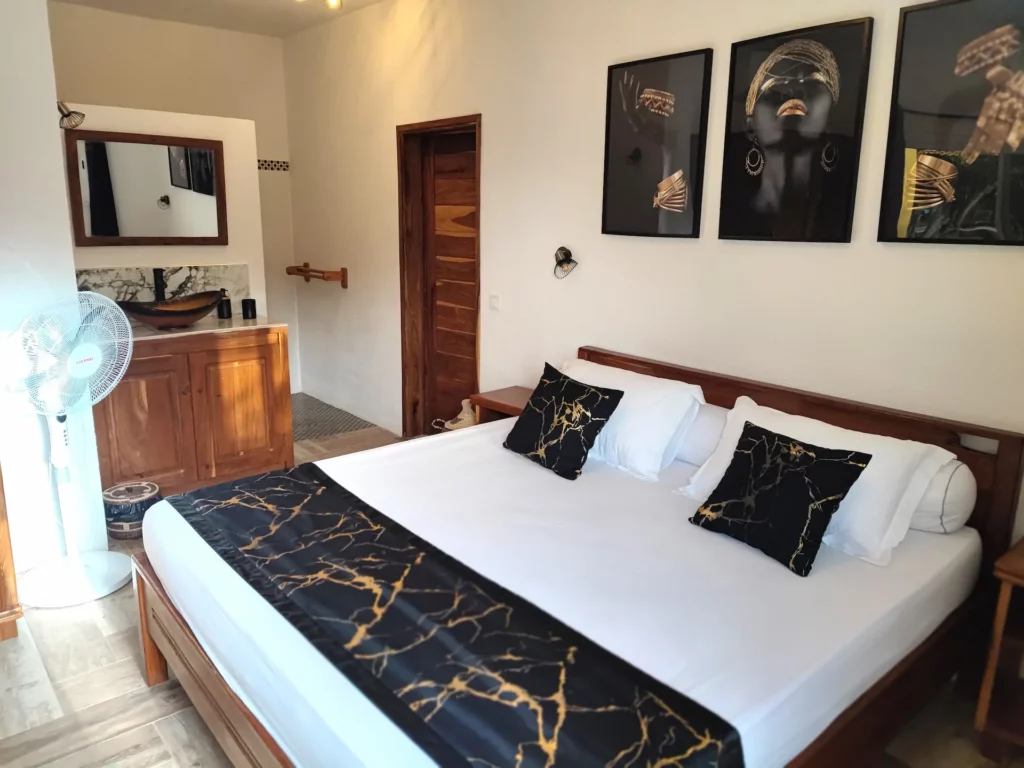 Le Campement Houéfa compte deux villas, avec chacune 3 chambres. Photo de la chambre numéro 5 avec son lit King size drapé de blanc, d'un chemin de lit et de coussins d'apparat de couleur noir et or, rappelant avec élégance les tons de l'espace douche et du triptyque surplombant le lit.